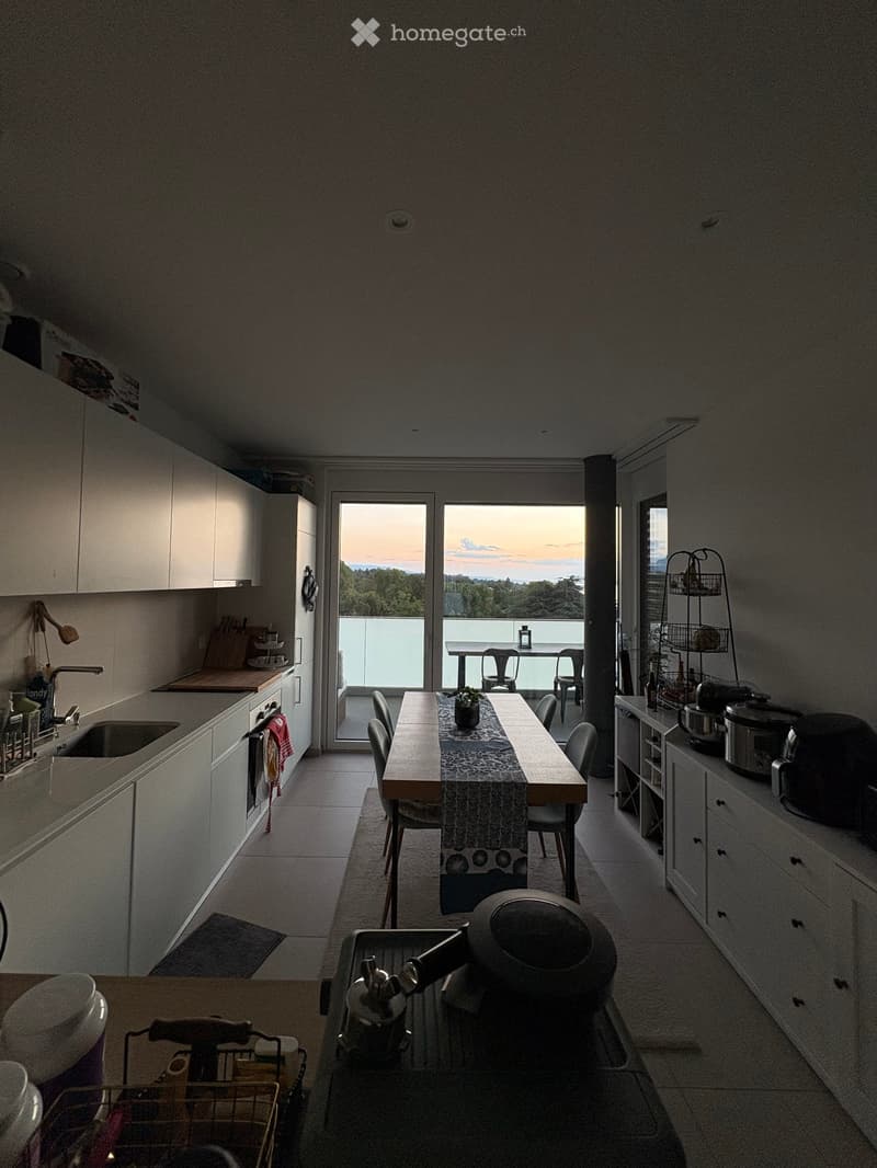 Modern kitchen with white cabinets, a central dining table, and a view of the sunset through large windows.