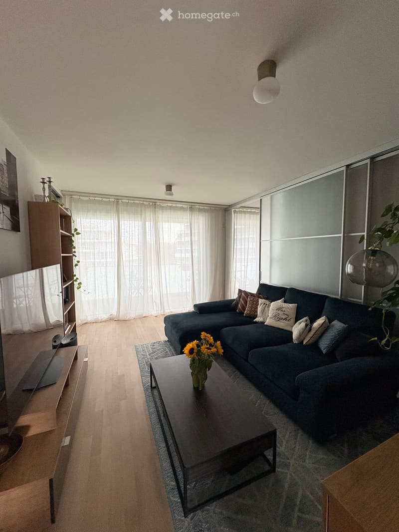 Modern living room with a dark blue sectional sofa, coffee table with a flower vase, bookshelf, large windows with curtains, and wall and table lamps.