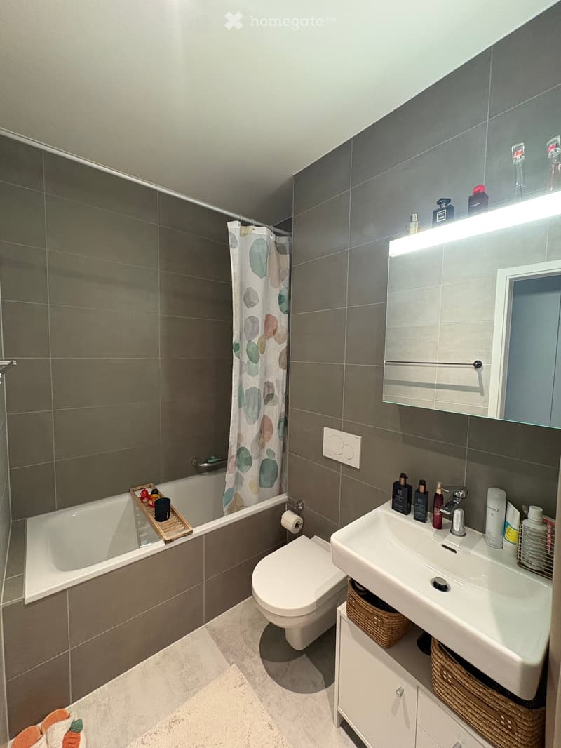 Modern bathroom with dark grey tiles, white bathtub, toilet, sink, mirror, and various toiletries. Features a shower curtain with a colorful abstract design and a wicker basket under the sink.