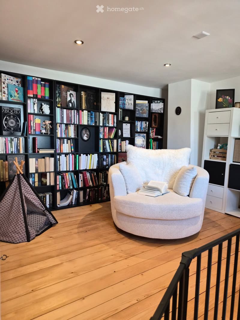 A cozy living room with a round white sofa, a large bookshelf filled with books, and a small storage cabinet. The room features wooden flooring and a staircase railing visible in the foreground.