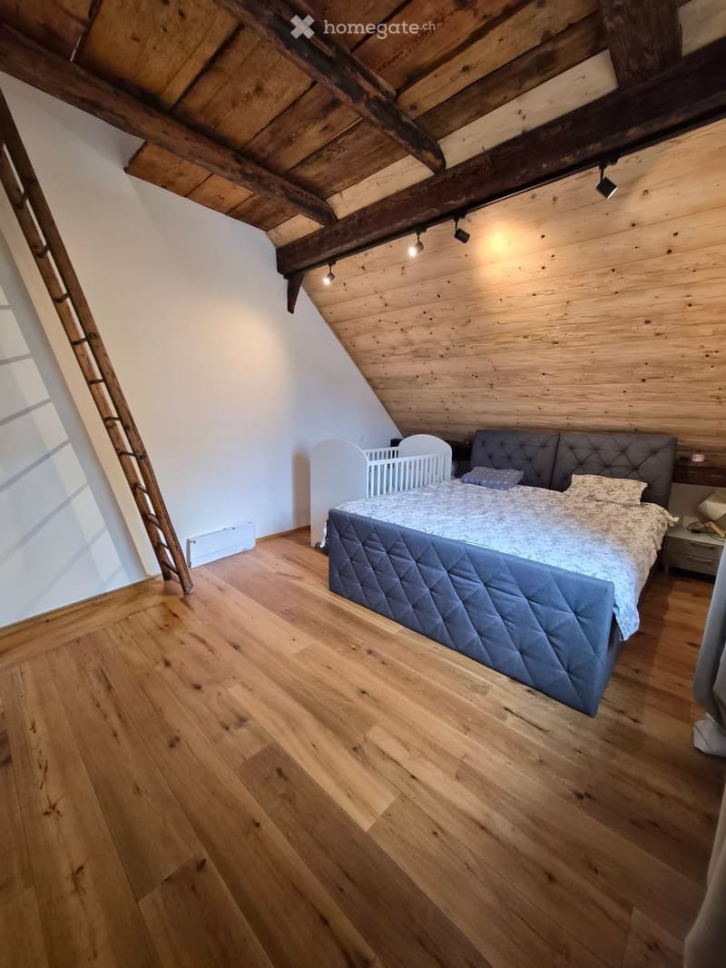Cozy attic bedroom with wooden floors, slanted wooden ceiling beams, and exposed wooden walls. Features a large bed with a quilted duvet, a small nightstand, and a white cradle.