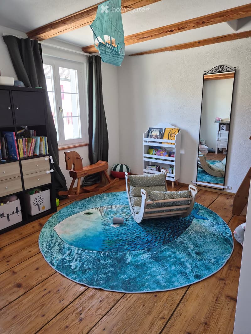 Child's room with wooden floor, large round rug, rocking horse, baby chair, bookshelf, mirror, and storage cabinet.