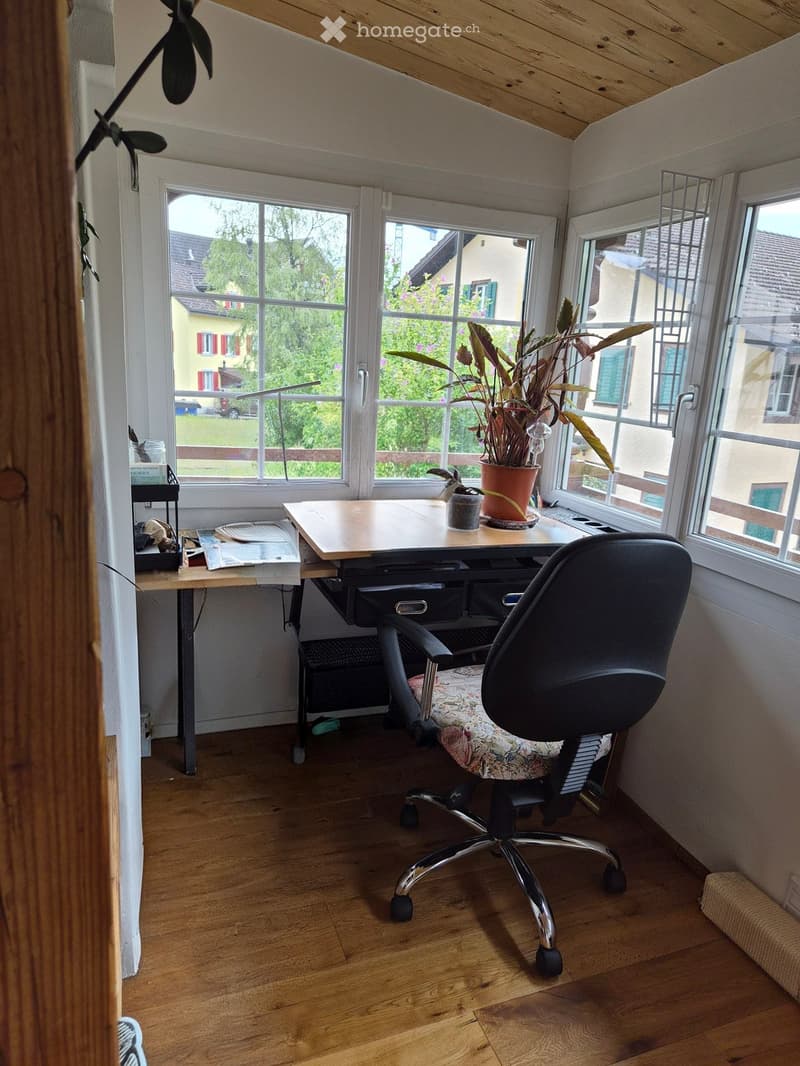 A small, well-lit room with wooden flooring and white walls, featuring a desk with a chair, a potted plant, and a lamp on the windowsill.