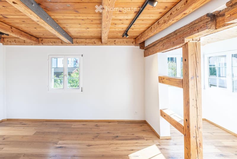 Empty room with wooden floor, white walls, exposed wooden beams, and large windows offering a view of greenery.