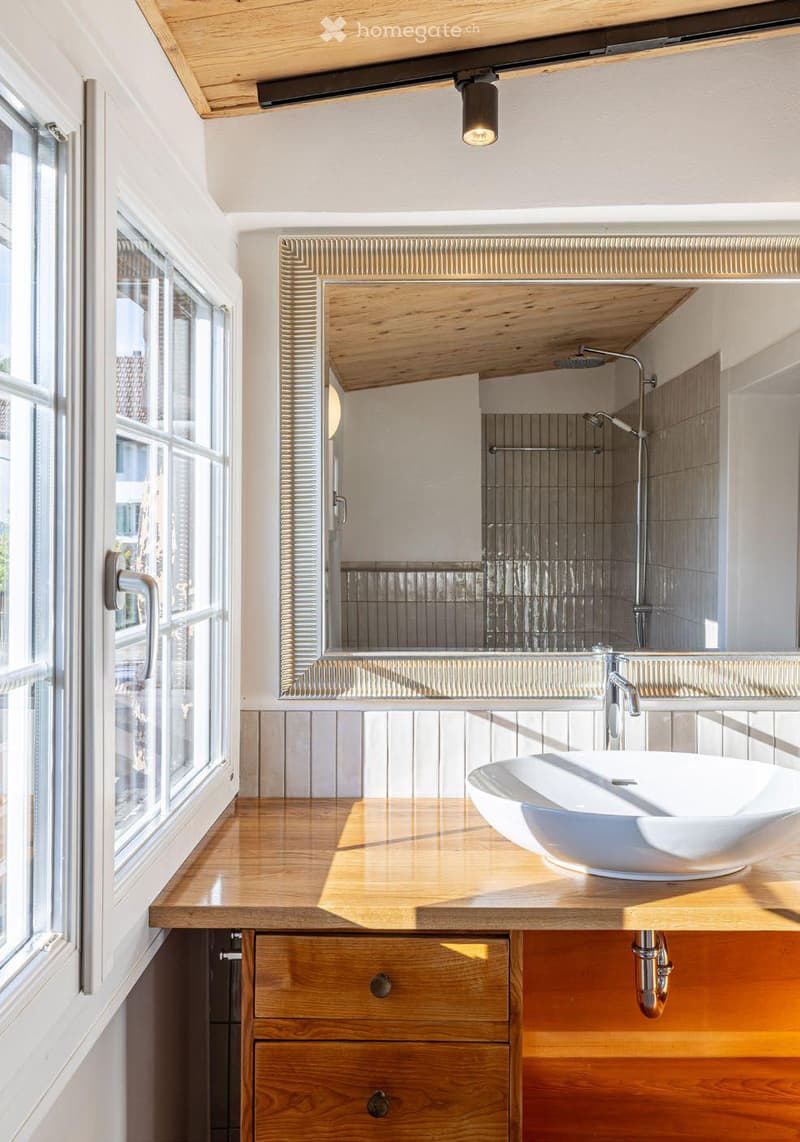 Modern bathroom with a wooden vanity, white sink, large mirror, and a shower area visible in the background.