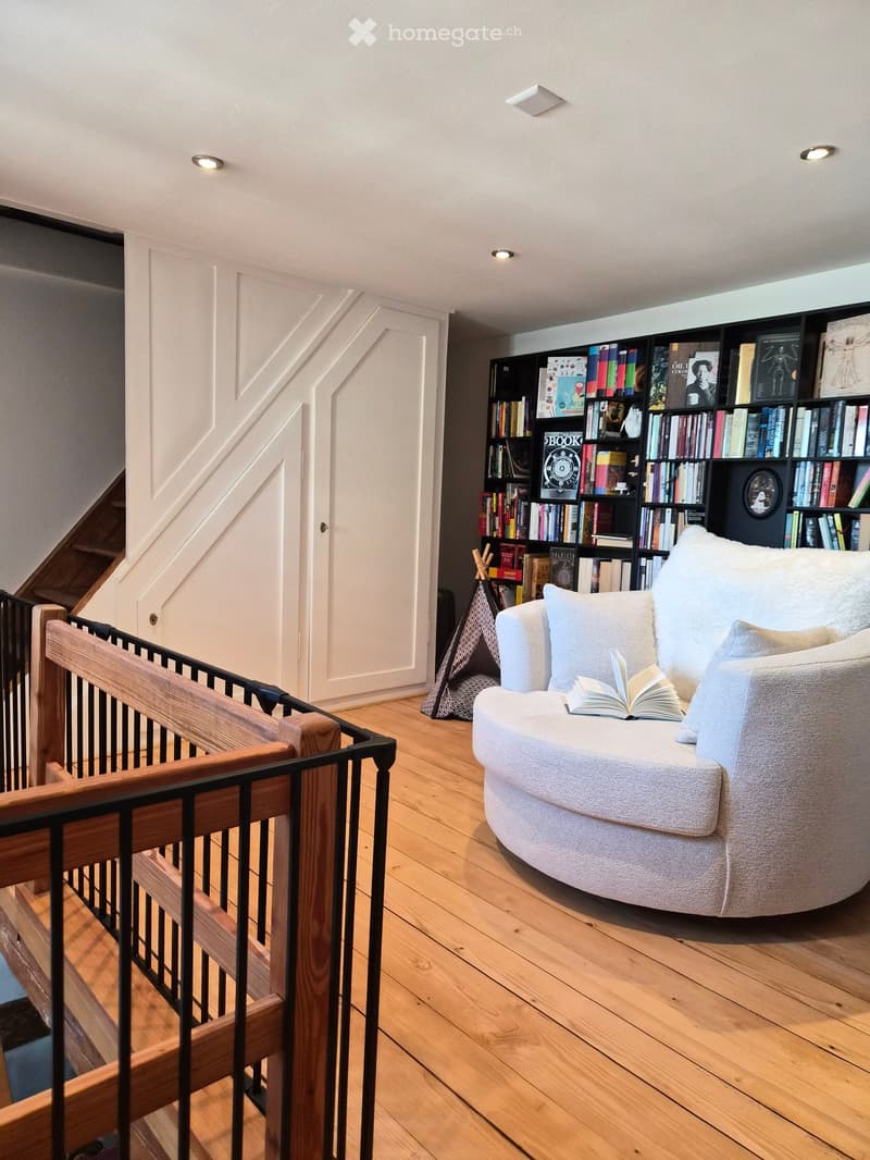 A cozy, well-lit room with wooden flooring, a round white sofa with a book resting on it, and a large black bookcase filled with books. A staircase with wooden railings is visible on the left side.