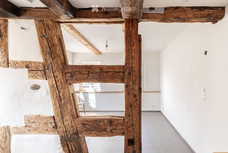 A modern hallway with exposed wooden beams and white walls, featuring a small seating area with a chair and table in the background.