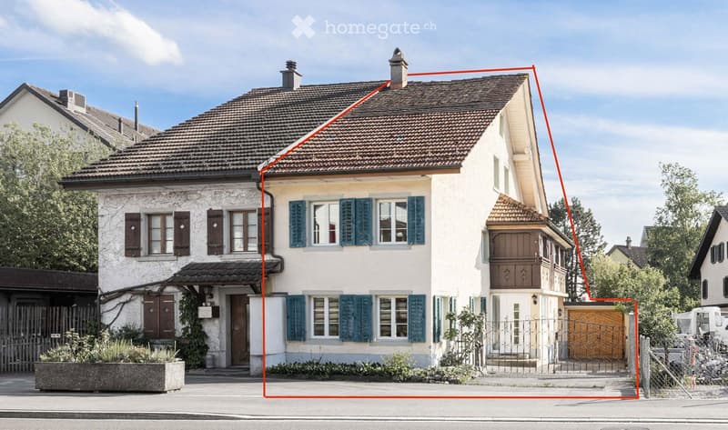 Two-story house with a tiled roof, white exterior, and blue shutters on windows. Features a small balcony on the right side and a wooden garage.