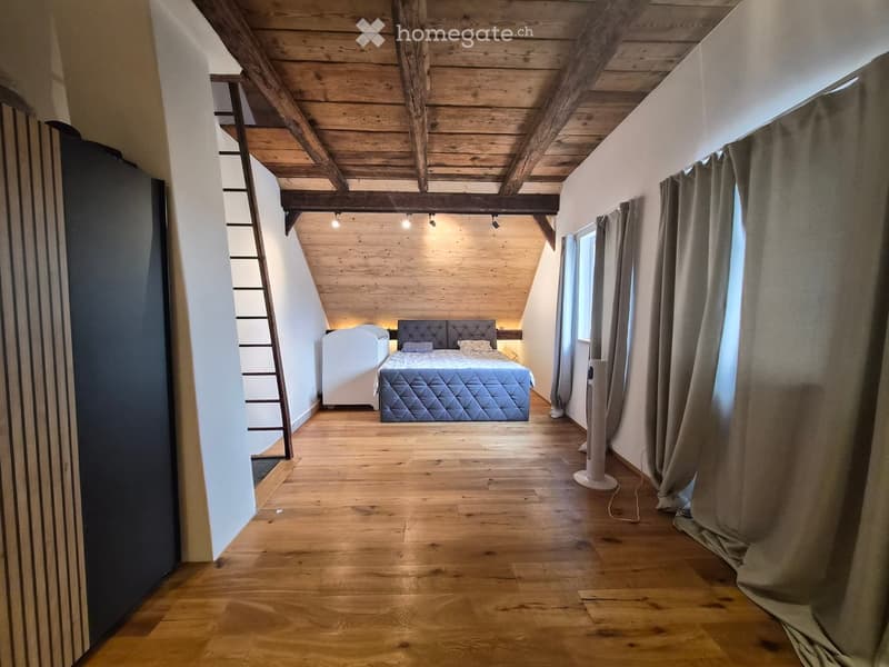 Cozy attic bedroom with wooden beams, exposed wooden ceiling, and wooden flooring. Features a bed with a quilted duvet, a nightstand, and a fan near the window.