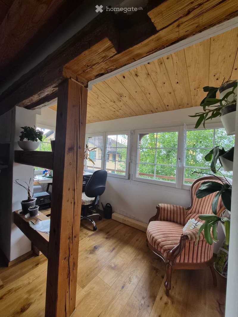 Cozy attic room with wooden beams, slanted wooden ceiling, large windows, a pink armchair, a small table with plants, and an office chair near a desk.