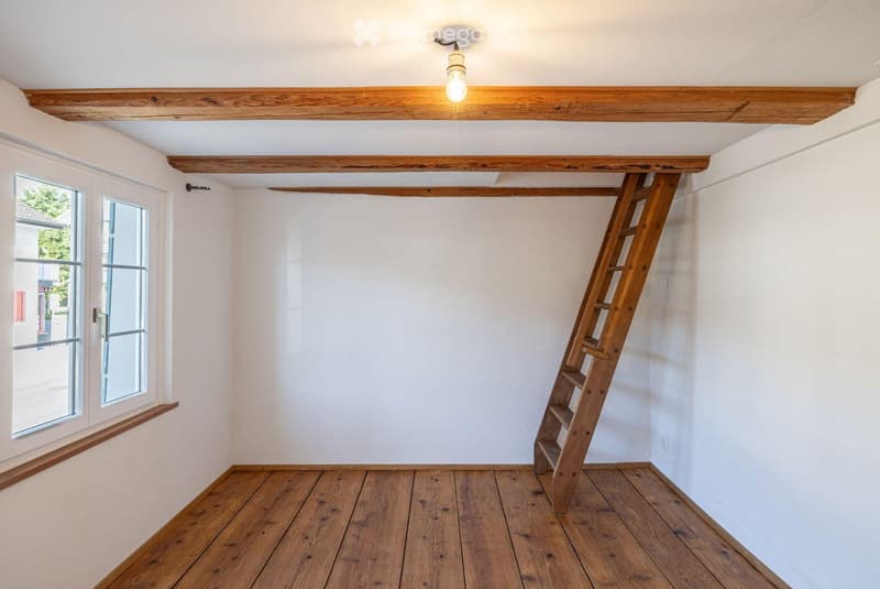 Empty room with wooden floor, white walls, exposed wooden beams, and a wooden ladder leading to an attic space. There is a window with a view of the outside.