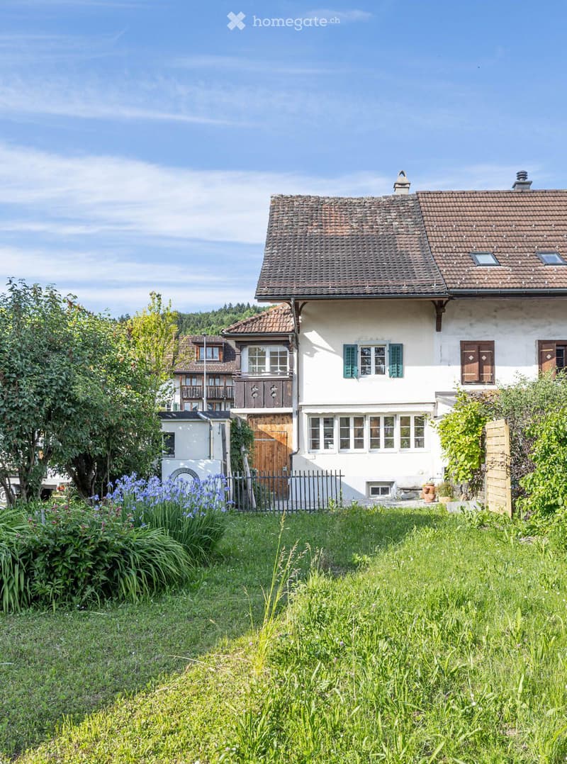 Two-story traditional house with white walls, brown tiled roof, multiple windows with shutters, and a fenced garden with flowers and greenery.