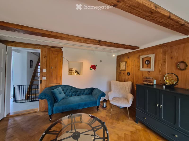 Living room with wooden ceiling beams, wooden walls, a blue sofa, a white armchair, a glass coffee table, a dark cabinet, and a mirror on the wall.