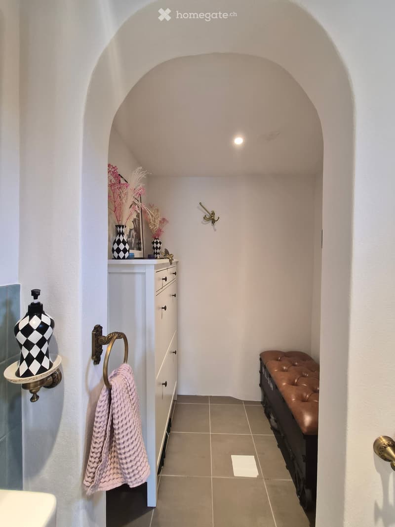 A small, well-lit hallway with a white arched ceiling, a small cabinet with drawers, a decorative vase, a picture frame, a pink towel, and a brown leather stool.