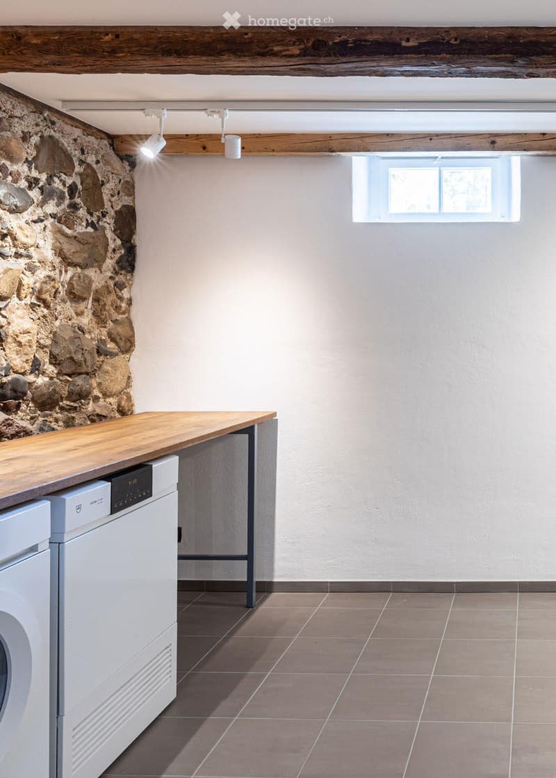 A modern, minimalist laundry room with a stone accent wall, wooden counter, and built-in appliances including a washing machine and dishwasher.