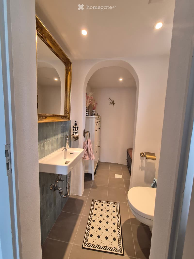 Elegant bathroom with a white sink, chrome faucet, large ornate mirror, toilet, and a patterned black and white rug. The walls are white with a decorative arched doorway leading to another space.
