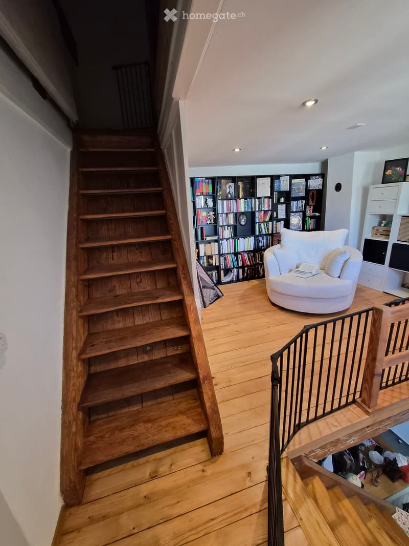 A cozy, well-lit room with wooden flooring, a round white sofa, a bookshelf filled with books, and a staircase leading upwards.