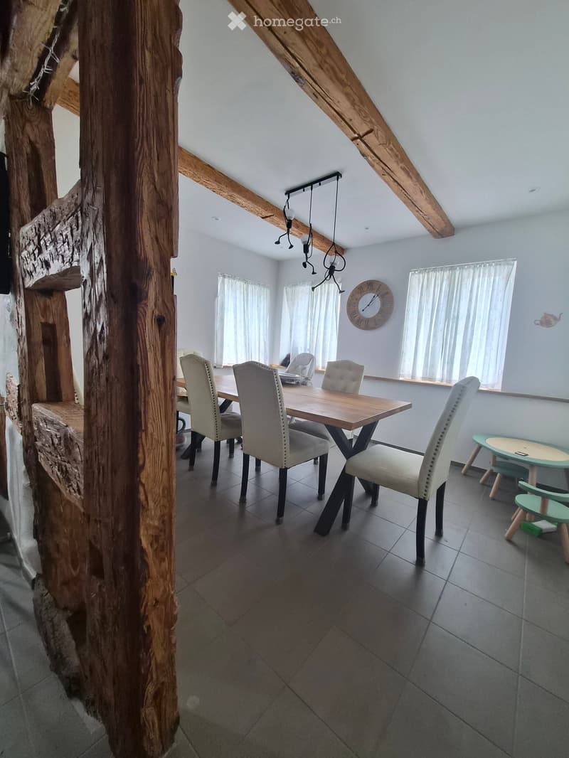 Dining room with a wooden dining table, four chairs, a clock on the wall, and a small green stool near the window.
