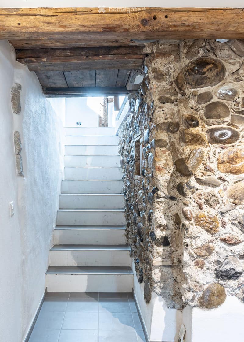 A narrow hallway with a stone wall on one side and a wooden ceiling. The floor is tiled, and a staircase leads upwards.