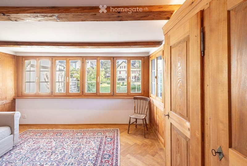 Cozy room with wooden beams, large windows, a sofa, a wooden chair, and a patterned rug on a wooden floor.