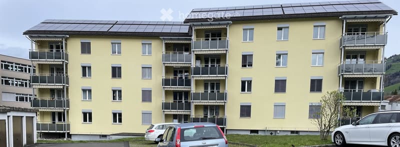 Multi-story yellow apartment building with multiple balconies, solar panels on the roof, and several parked cars in front.