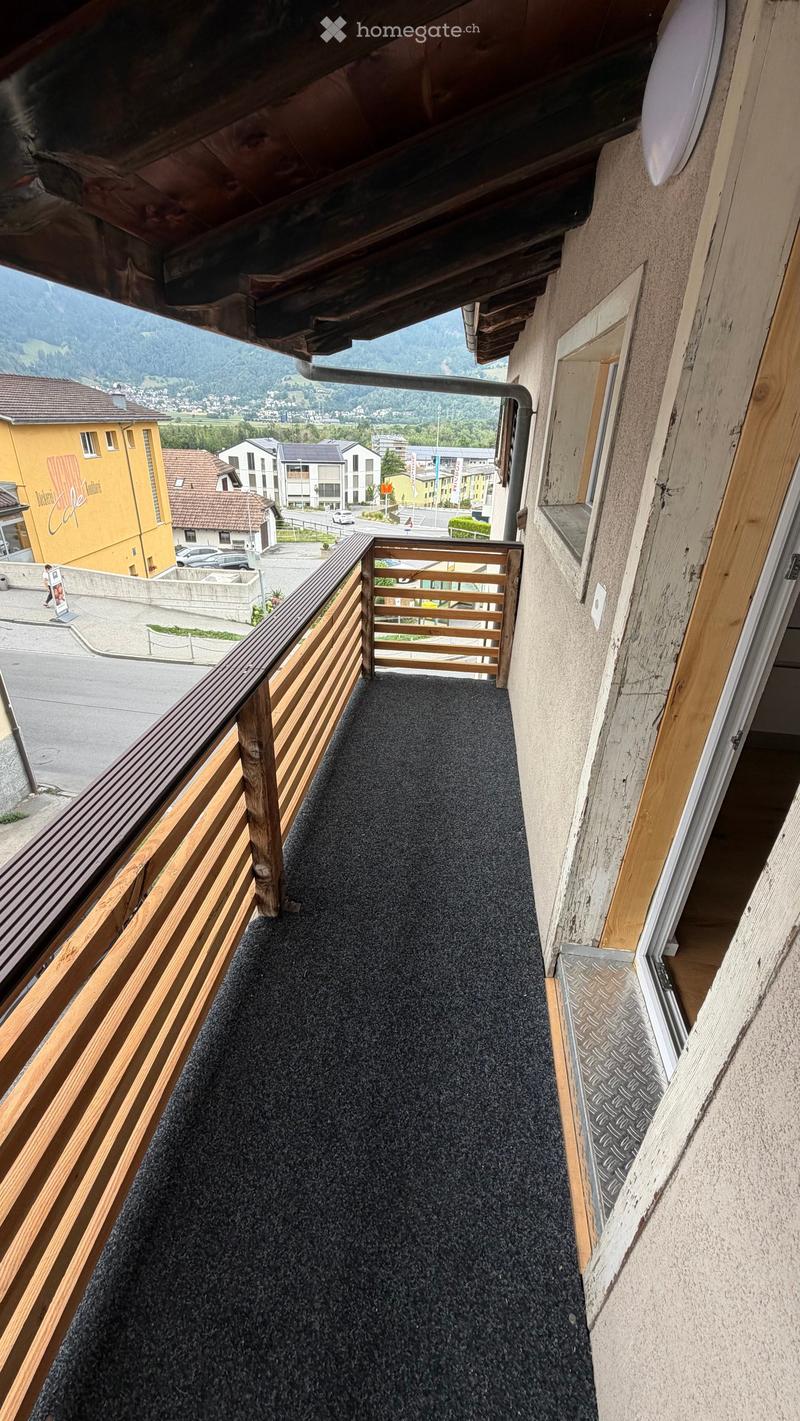 Balcony with wooden railing, carpeted floor, overlooking a town with buildings and mountains in the background.