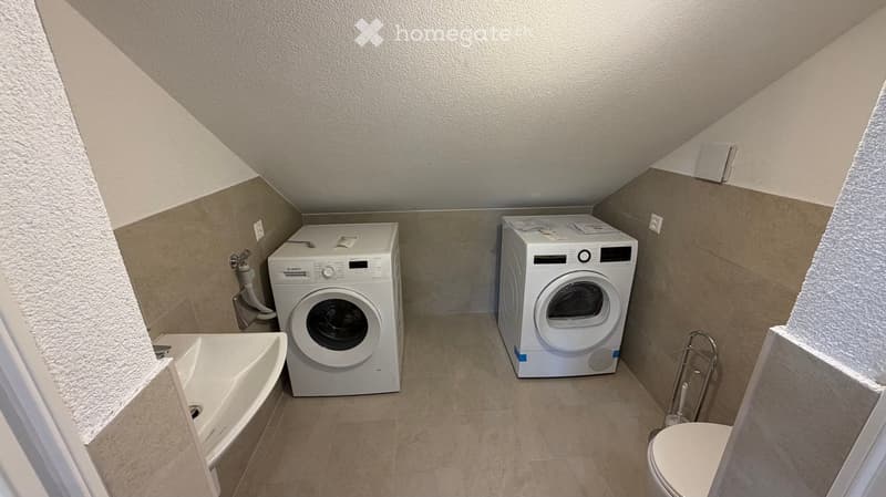 Compact bathroom with a washing machine, dryer, bathtub, and toilet, featuring tiled walls and a slanted ceiling.