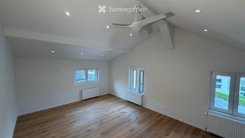 Empty room with wooden flooring, white walls, and multiple windows, including a large window with a view of the street.