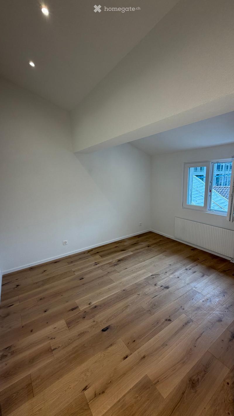 Empty room with wooden flooring, white walls, and a window with a view of a rooftop area.
