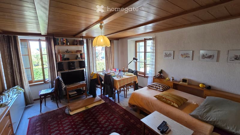Cozy bedroom with wooden ceiling, bed, chair, table, shelf, lamp, and artwork on the wall.