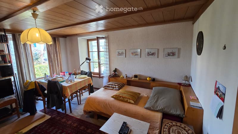 Cozy bedroom with a double bed, bedside table, desk, and a hanging lamp. The room features wooden ceilings, large windows with curtains, and various decorative items.