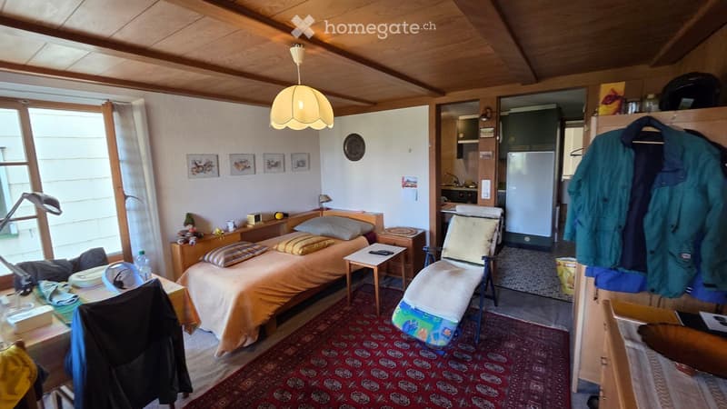 Cozy attic bedroom with wooden beams, two single beds, a chair, a small table, a lamp, and a small kitchen area with a refrigerator and cabinets.