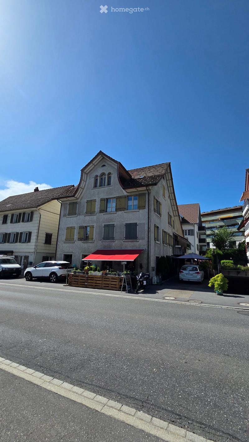 Three-story building with a red awning, multiple windows, and parked cars in front.