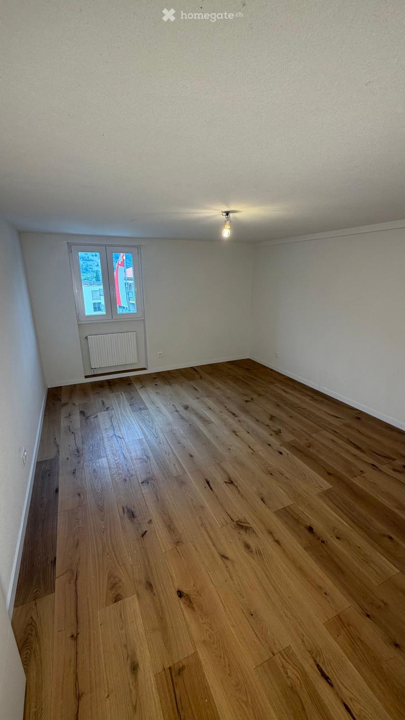 Empty room with light-colored wooden flooring, white walls, and a single window with a radiator below it.