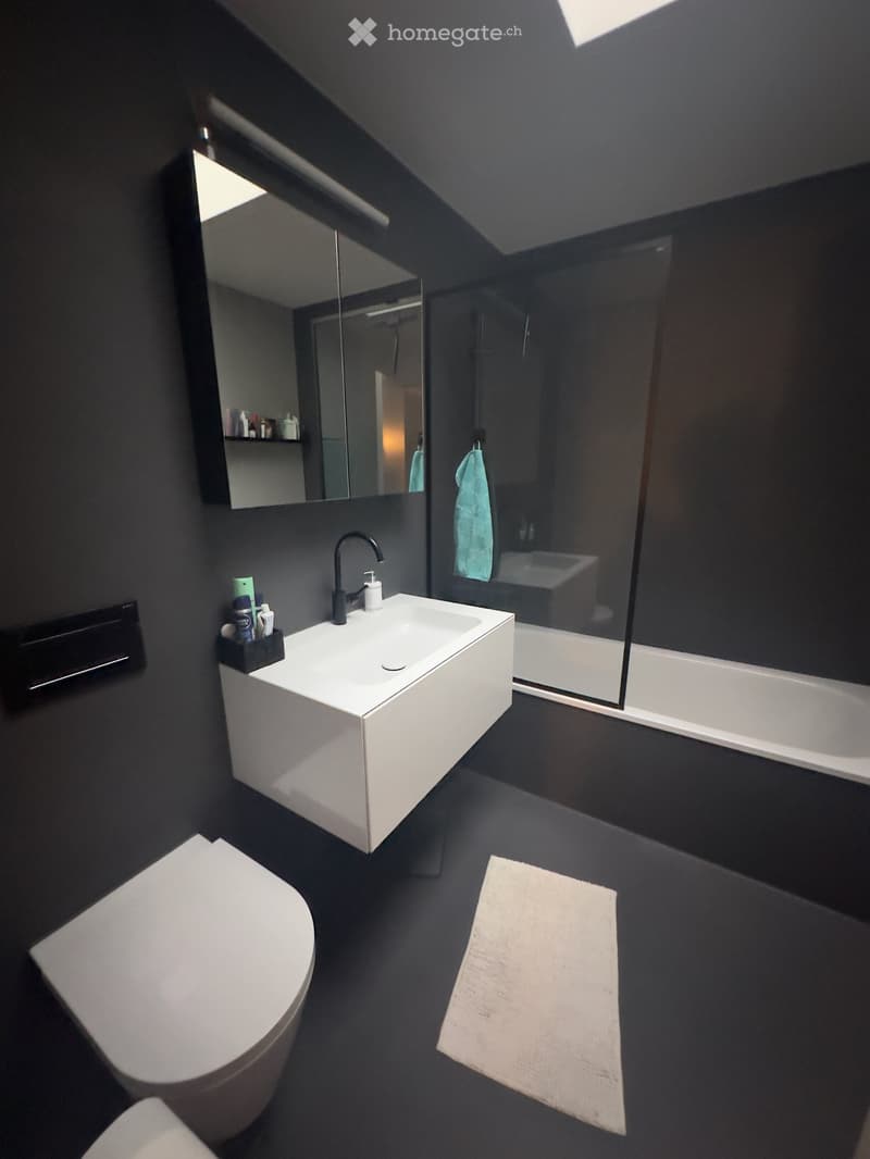 Modern bathroom with dark walls and floor, white sink, toilet, and bathtub, featuring a large mirror and a towel on the floor.