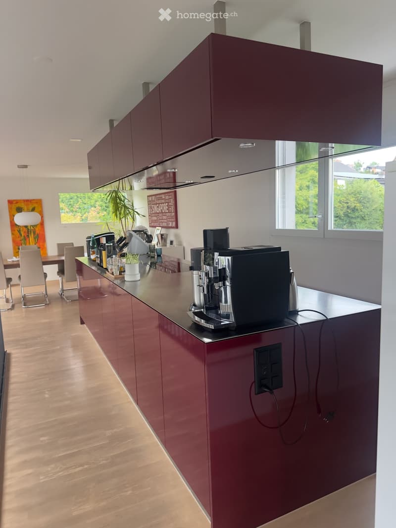 Modern kitchen with dark red cabinets, stainless steel appliances, and a central island. The island features a coffee machine, various bottles, and a small plant. The background shows a dining area with chairs and a table.