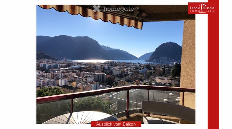 Balcony with a panoramic view of a cityscape, lake, and mountains under a clear blue sky.