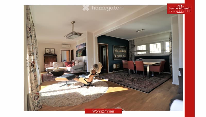 A spacious, well-lit living room with a modern design. Features include a white sofa, coffee table, dining table with chairs, a cabinet, picture frames, a large window with curtains, and a floor lamp. The room has wooden flooring and a cozy, inviting atmo
