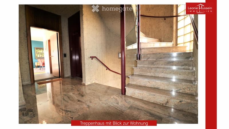Spacious hallway with polished marble floors, a staircase with a polished metal railing, and an open doorway leading to a room with a red chair and a painting on the wall.