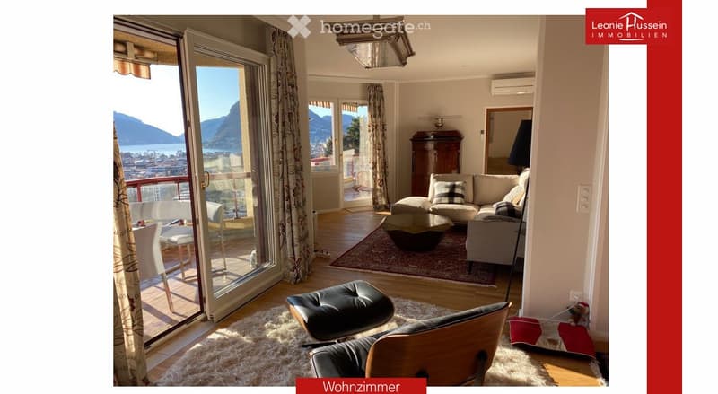 Bright living room with large windows and a view of the lake and mountains, featuring a sofa, coffee table, ottoman, chairs, and a dining area with a table and chairs.