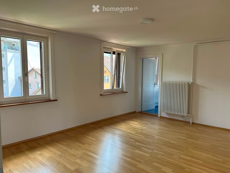 Empty room with wooden flooring, white walls, large windows, and a radiator. No furniture or equipment present.