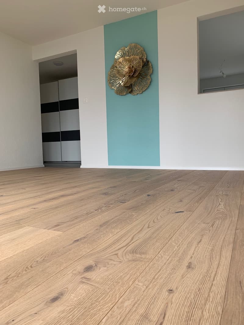 Empty room with light wooden flooring, white walls, and a decorative gold flower wall piece. A closet with black and white doors is visible in the background.