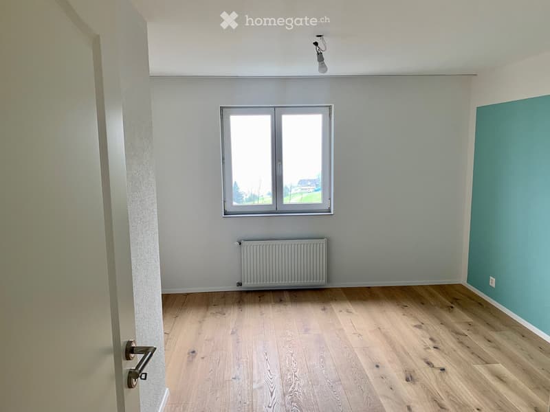 Empty room with light wooden flooring, white walls, a window with double panes, a radiator, and a door with a brass handle.