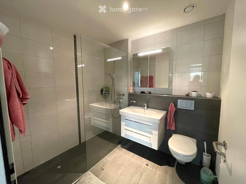 Modern bathroom with a bathtub, sink, toilet, mirror, and a glass-enclosed shower area. The sink has a modern faucet and a white cabinet with drawers. A towel is draped on the side of the sink, and a toilet brush is visible near the toilet.