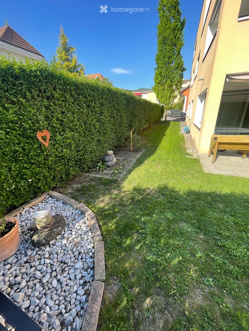 Well-maintained garden with a gravel pathway, neatly trimmed hedges, a small stone water feature, and a wooden bench near the house.