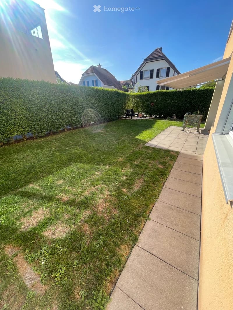 Well-maintained garden with a lawn, trimmed hedges, a concrete pathway, and a small seating area with a bench and a table.