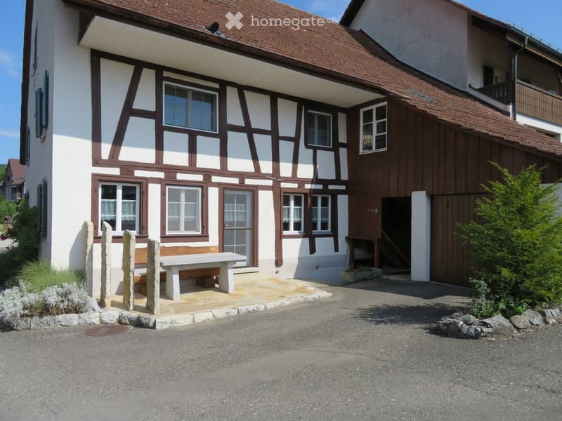 Two-story house with white and brown timber framing, multiple windows, and a stone bench in the front yard.
