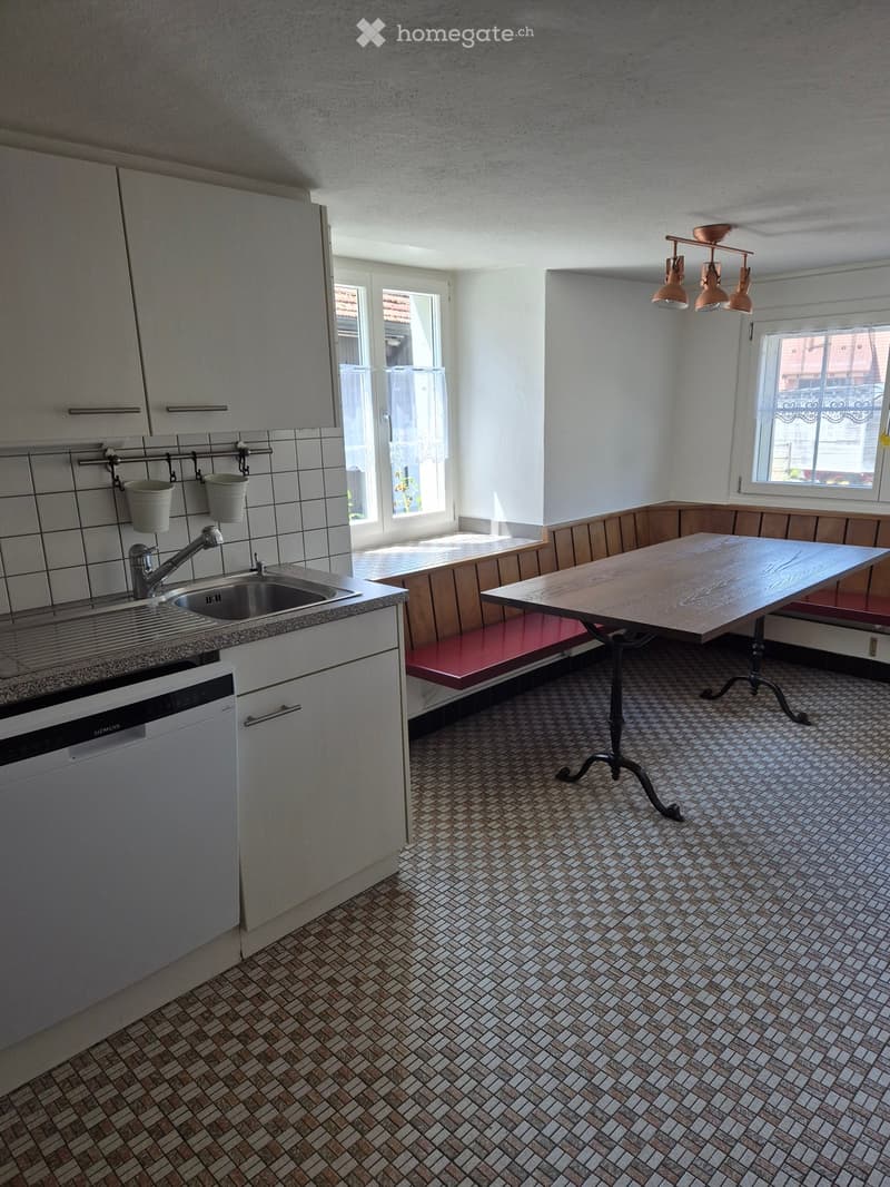 Bright kitchen with tiled walls, a sink, a dishwasher, a wooden table with a red bench, and pendant lights.