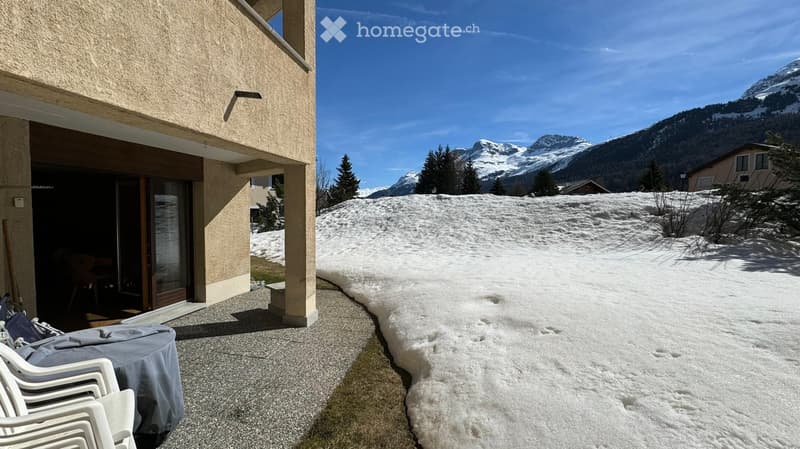 Balcony with white plastic chairs and a covered table, overlooking a snowy landscape with mountains in the background.