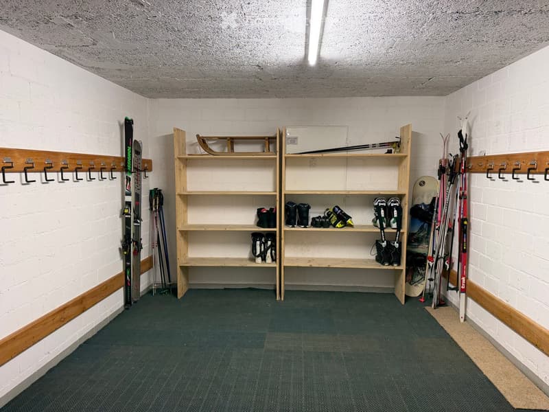 A small, organized storage area with white brick walls, wooden shelves, and hooks holding skis, ski poles, and snowboards. The floor is green, and the ceiling has exposed lighting.